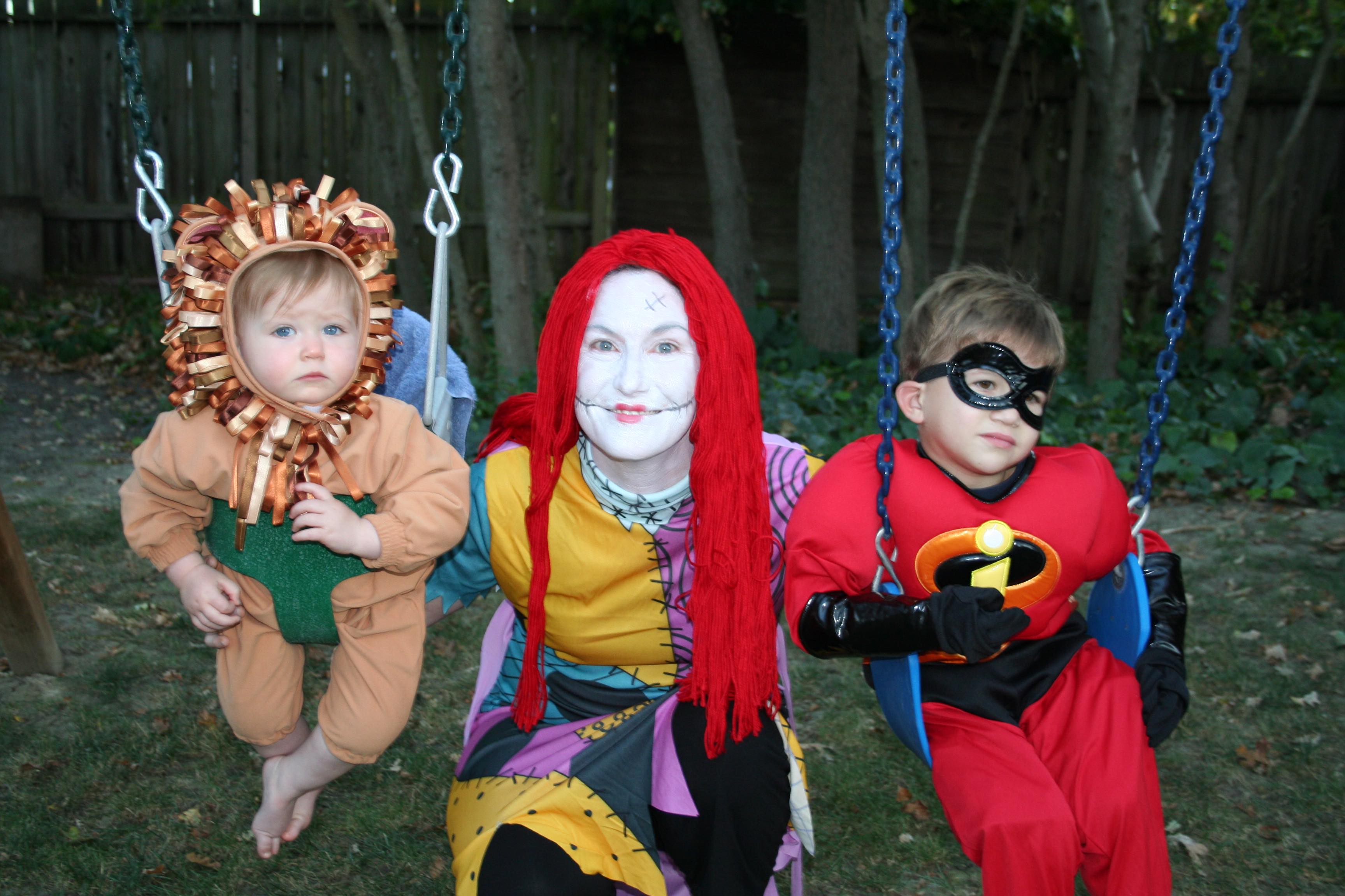 Three children in Halloween costumes on backyard swings — a lion, Sally from The Nightmare Before Christmas, and a superhero