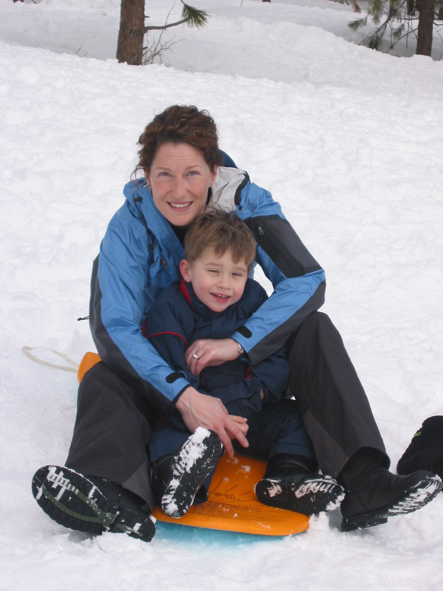 A mother hugging her young son on an orange sled in fresh snow among pine trees